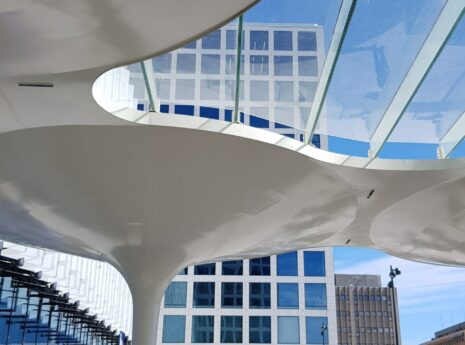 Moderni arkkitehtoninen rakenne, jossa on suuret, valkoiset, kaarevat tukirakenteet ja lasikatto. Taustalla näkyy rakennuksia, joissa on ruudukkomuotoinen ikkunajärjestely.