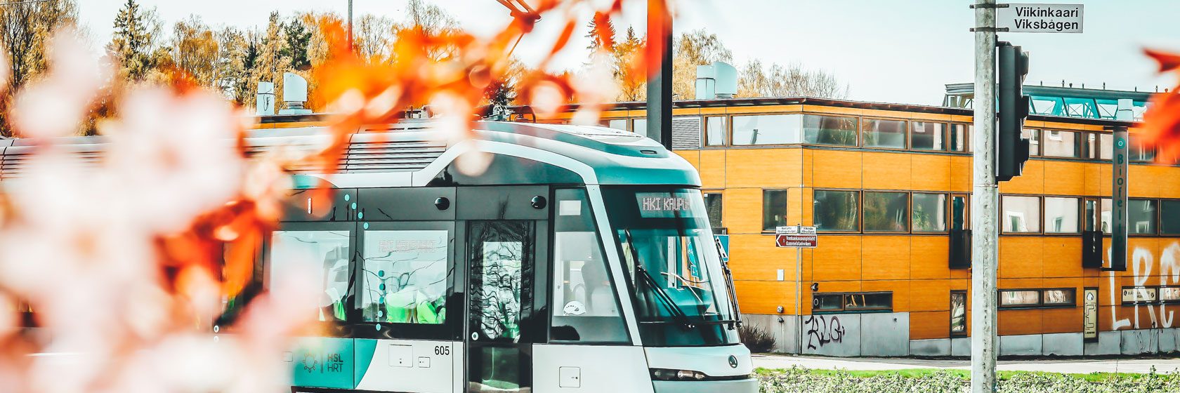 Raide-Jokeri raitiovaunu kesäisessä maisemassa, etualla kukkia
