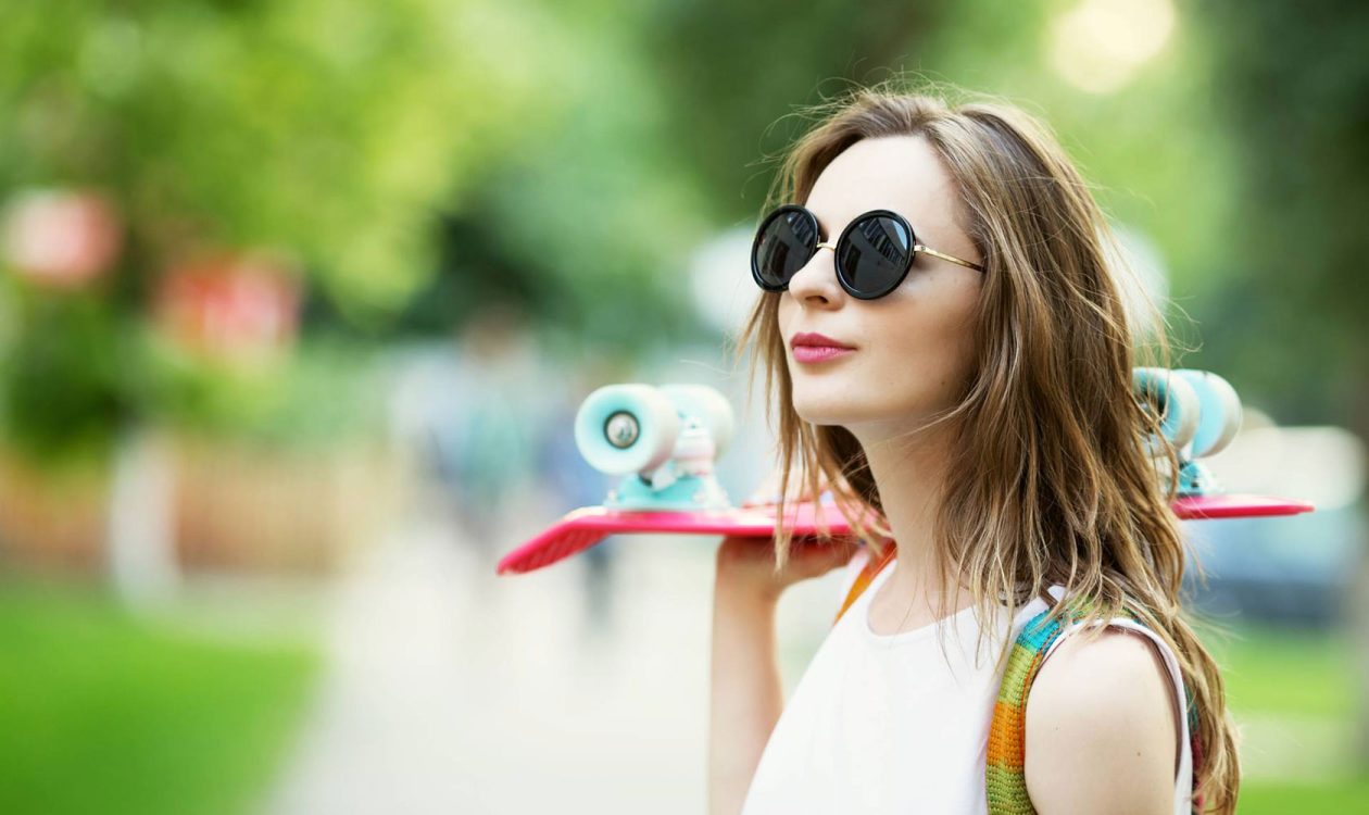 Tyttö puistossa skeittilauta olkapäällä ja taustalla puistomaisema.