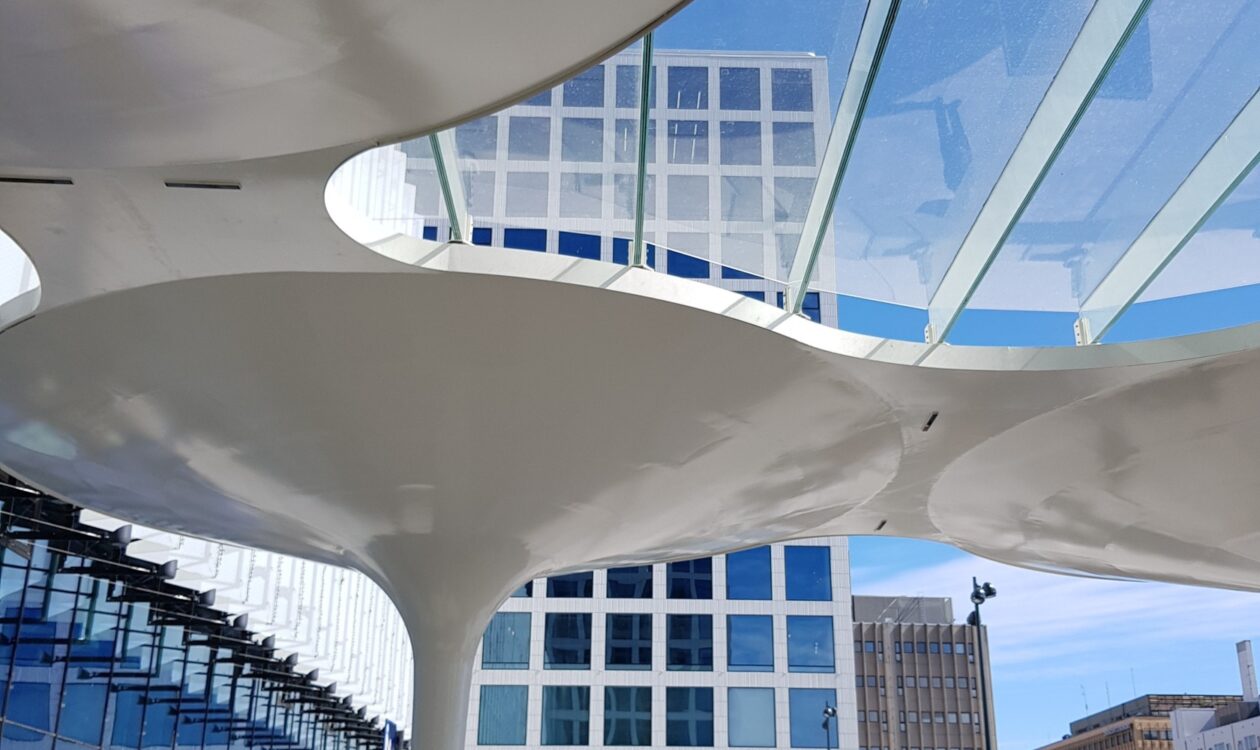 arkkitehtoninen rakenne, jossa on valkoisia kaarevia kattoelementtejä ja lasikatto. Taustalla näkyy korkea rakennus, jossa on sinisiä ja valkoisia ruutuikkunoita.