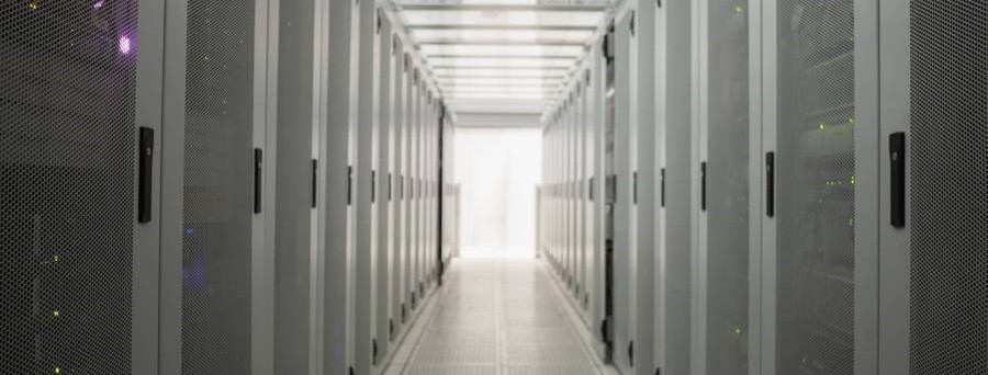 The image shows a long corridor with server cabinets lined up on both sides. At the end of the corridor, a bright light is visible. The server cabinets are gray with black handles.