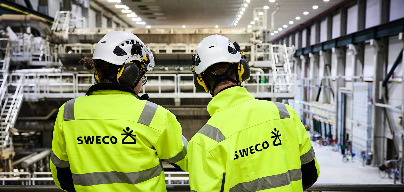 The image shows two individuals wearing high-visibility jackets and white helmets with ear protection, standing inside an industrial facility. The jackets have the text "SWECO" and a logo on the back. The facility appears to be large, with machinery and equipment in the background.