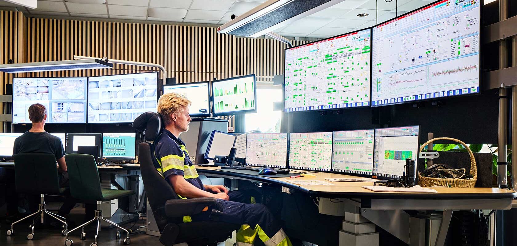 Valvontahuone, jossa on useita näyttöjä, jotka näyttävät erilaisia tietoja ja kaavioita. Kaksi henkilöä istuu työpöytien ääressä, kumpikin työskentelee tietokoneilla, joiden edessä on useita näyttöjä.