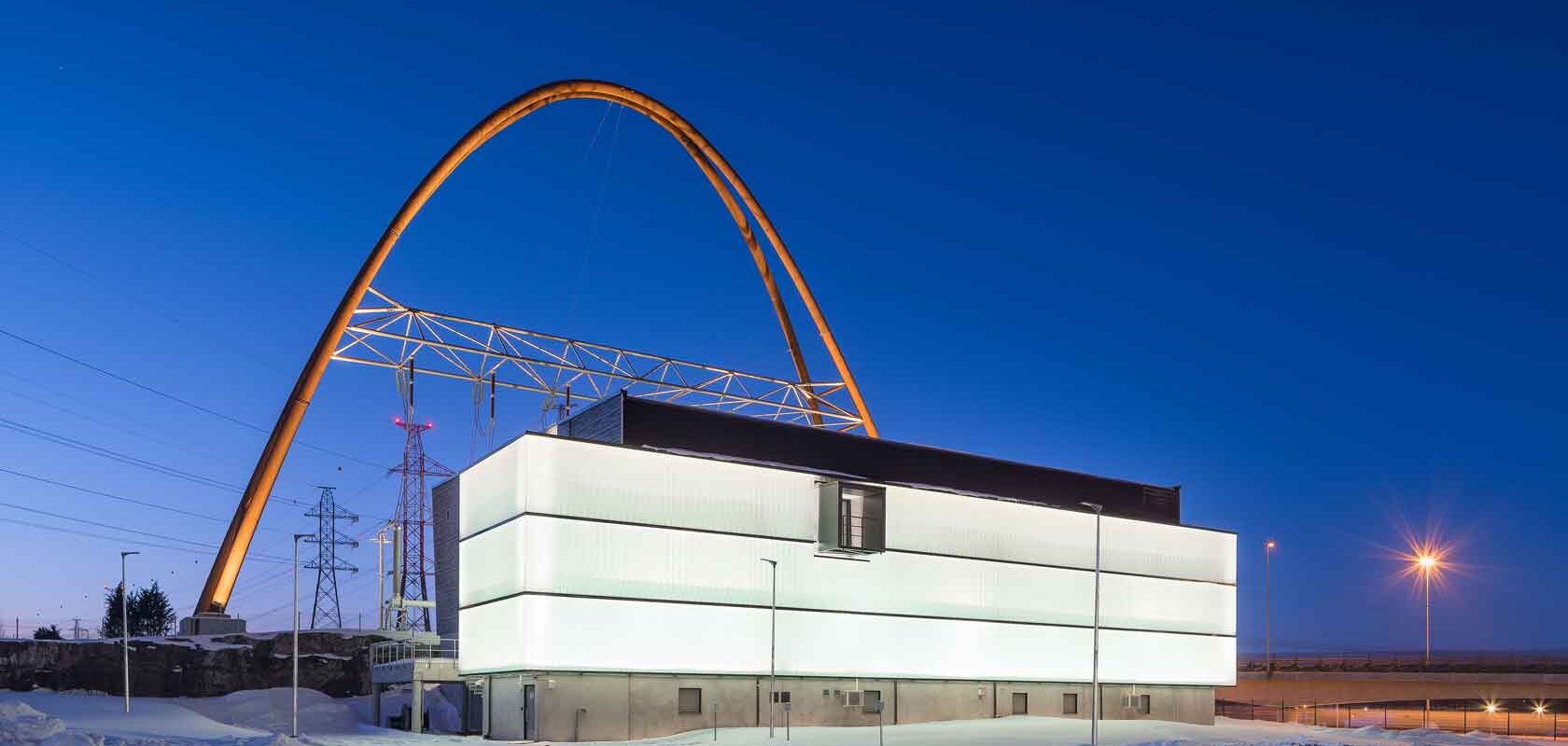 Kuvassa Länsisalmen sähköasema. Moderni rakennus, jossa on hehkuva julkisivu, sijaitsee lumisessa maisemassa. Taustalla näkyy suuri keltainen kaarirakenne ja useita sähkölinjoja ja torneja.