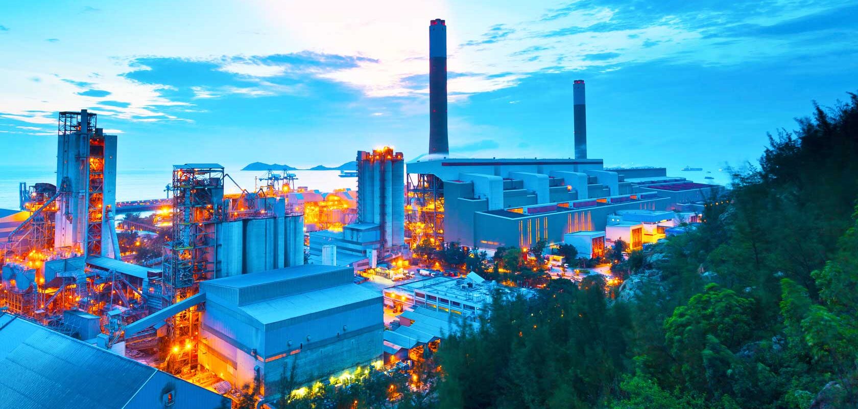 An industrial area with several large buildings and smokestacks. The area is brightly lit, with numerous lights reflecting off the surrounding landscape. In the background, the sky features clouds and colors from either a sunset or sunrise.