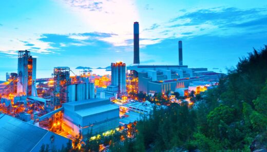 An industrial area with several large buildings and smokestacks. The area is brightly lit, with numerous lights reflecting off the surrounding landscape. In the background, the sky features clouds and colors from either a sunset or sunrise.