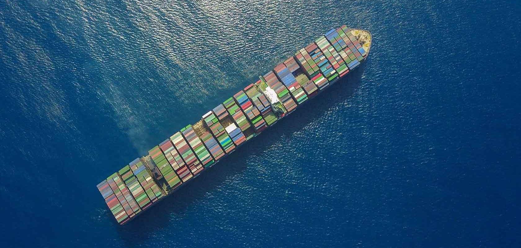 Aerial view of a large cargo ship sailing in the ocean, loaded with colorful shipping containers arranged in rows on its deck. The surrounding water is deep blue, with no other visible objects or landmasses.