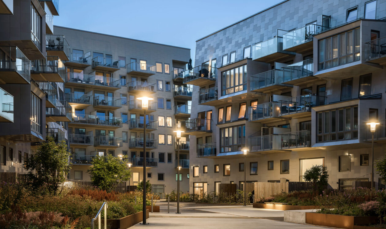 Moderni asuinkerrostalojen sisäpiha iltavalaistuksessa, jossa näkyy lasikaiteisia parvekkeita, vehreää piha-aluetta ja tunnelmallisia katuvaloja.