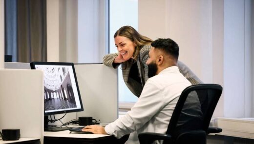 Two people are in an office setting, looking at a computer screen. The person sitting is wearing a white shirt and using the computer mouse, while the person standing is leaning over the partition to observe the screen