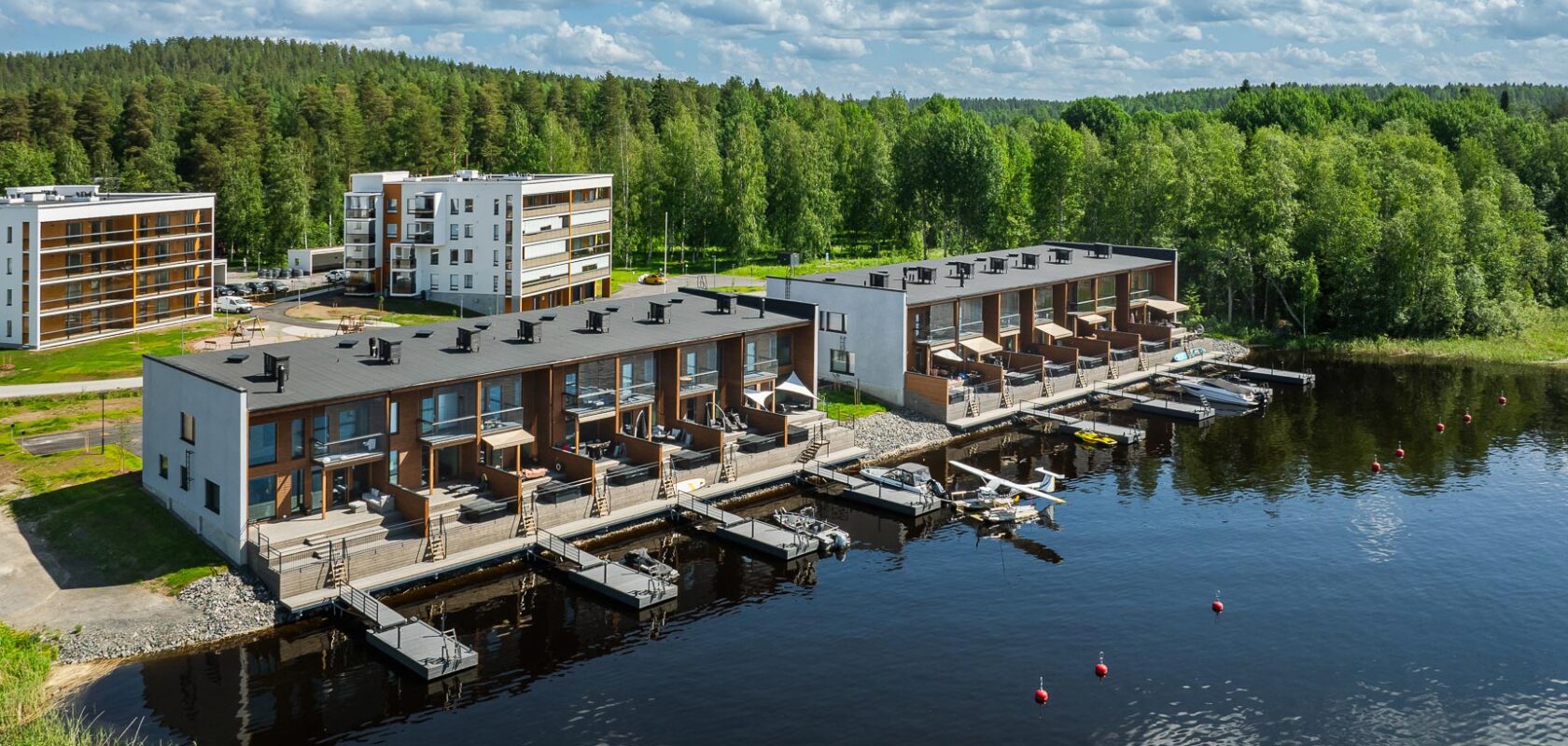Moderni asuinrakennus, joka sijaitsee vesistön, todennäköisesti järven tai joen, rannalla. Rakennuksissa on useita kerroksia ja parvekkeita, ja joissakin asunnoissa on suora pääsy laitureille.
