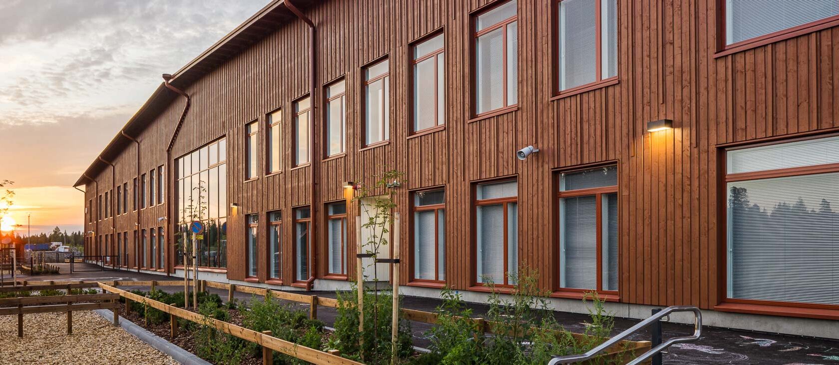Moderni puinen kaksikerroksinen rakennus, jossa on pystysuora puupaneelointi sekä punakehyksiset ikkunat. Rakennuksen edessä on pieniä puita ja kasveja, sekä puinen aita ja päällystetty kävelytie