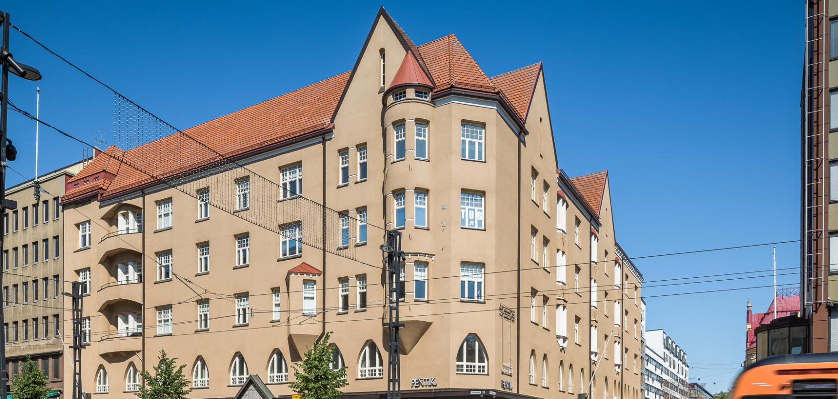 Katunäkymä suuresta, monikerroksisesta rakennuksesta, jossa on beige seinät ja punatiilikatot. Rakennuksessa on useita ikkunoita, joissakin pieniä parvekkeita.