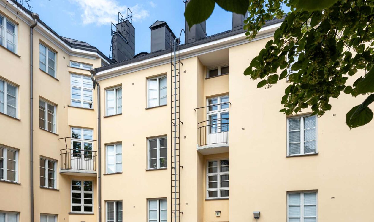 vaaleankeltaista kerrostaloa, jossa on useita parvekkeita ja ikkunoita. Talon keskellä on tikkaat, jotka ulottuvat katolle asti. Oikeassa reunassa näkyy puun oksia ja lehtiä.