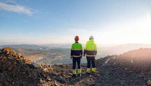 Two individuals wearing high-visibility work clothing and helmets standing on a rocky terrain, overlooking what appears to be a mining or construction site in the distance.