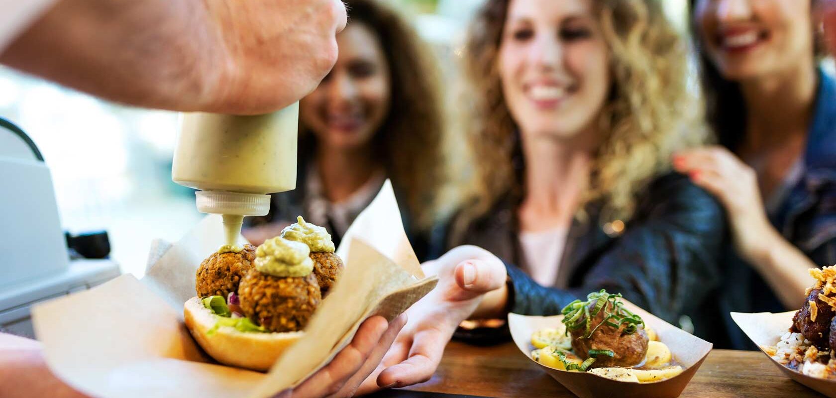 Henkilö puristaa kastiketta annokseen, joka sisältää falafeliä. Taustalla useita ihmisiä ruokakojulla tai ravintolan tiskillä.
