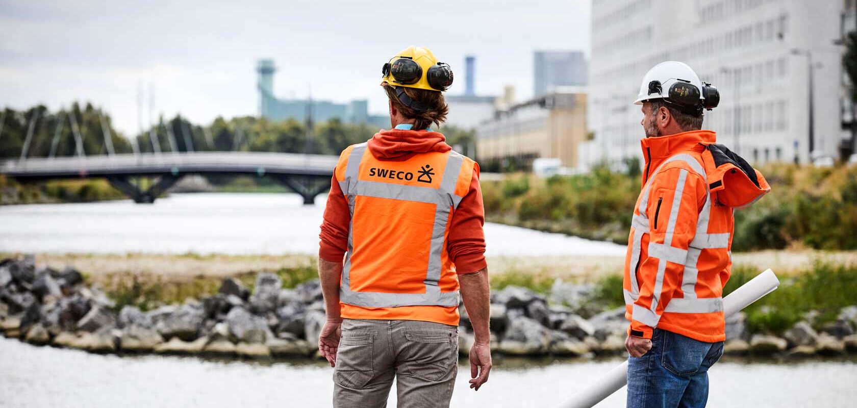 Kuvassa on kaksi henkilöä, jotka käyttävät oransseja huomiotakkeja, joissa on heijastavat raidat. Molemmat henkilöt käyttävät kypäriä ja näyttävät olevan rakennustyömaalla tai työmaalla veden äärellä.