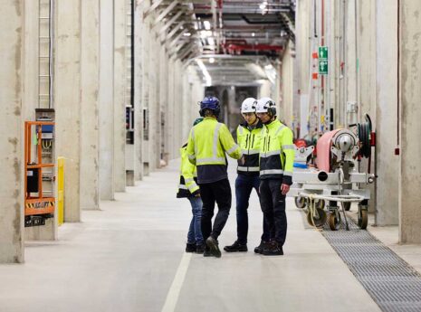Four workers wearing high-visibility jackets and helmets standing in a large industrial hallway. The hallway is lined with concrete pillars and various industrial equipment, including pipes and machinery.