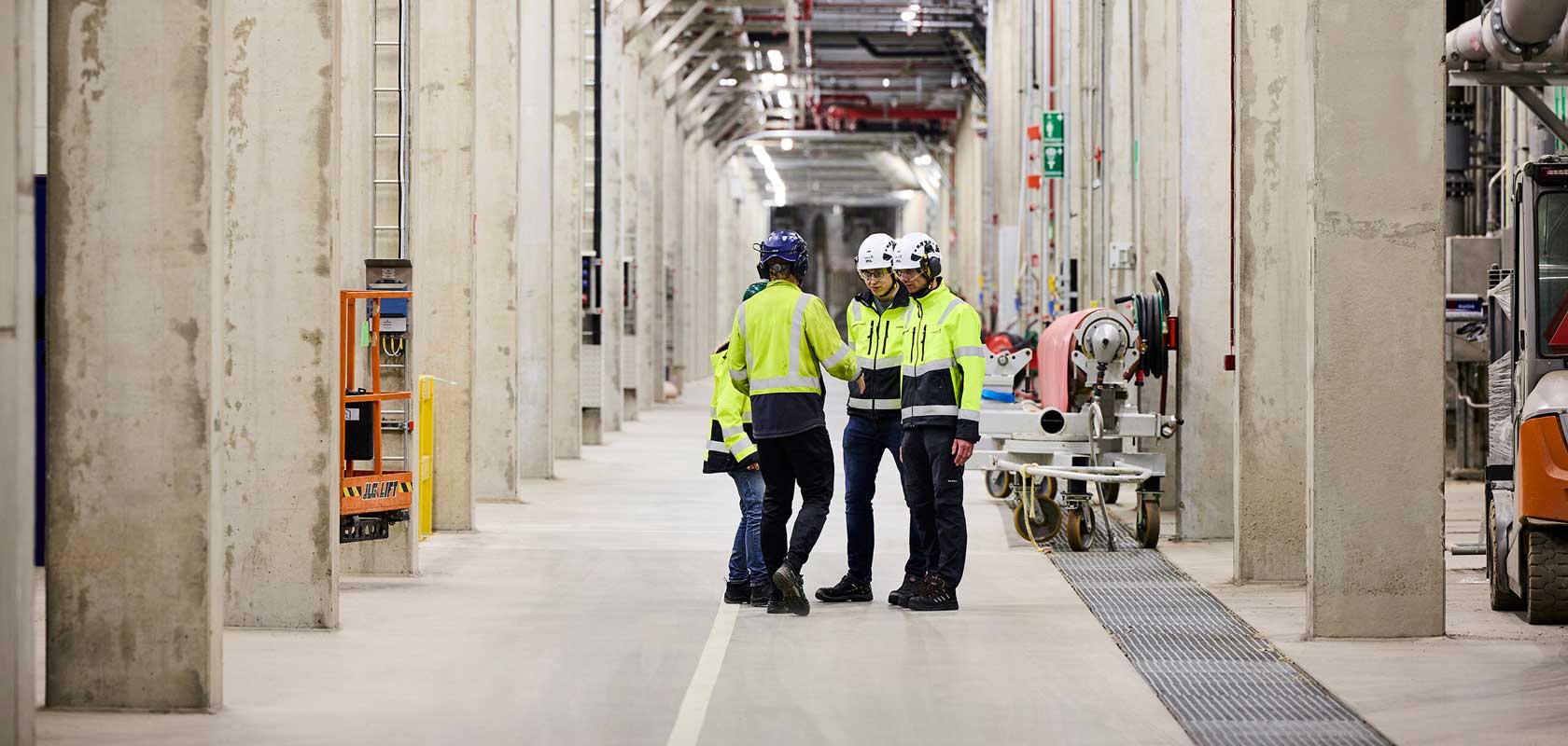 Four workers wearing high-visibility jackets and helmets standing in a large industrial hallway. The hallway is lined with concrete pillars and various industrial equipment, including pipes and machinery.