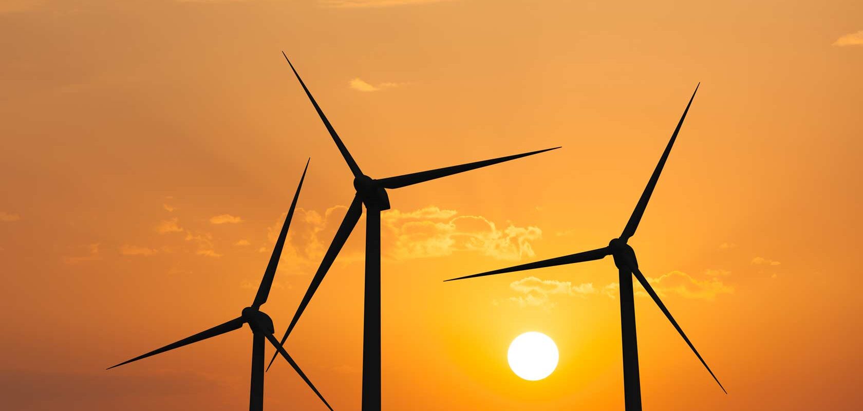 Kolme tuulivoimalaa siluettina oranssia auringonlaskua vasten. Aurinko näkyy lähellä horisonttia, luoden lämpimän hehkun ja rauhallisen tunnelman