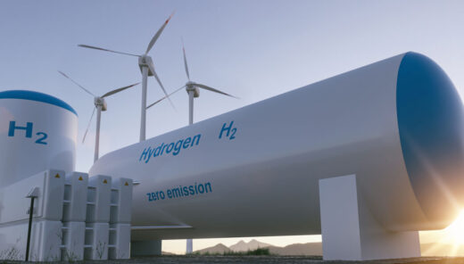 A large hydrogen storage tank labeled "Hydrogen H₂" and "zero emission" is situated outdoors with three wind turbines in the background. The tank has a cylindrical shape with rounded ends, and there are smaller structures nearby. The scene is set during sunset or sunrise, with mountains visible in the distance.