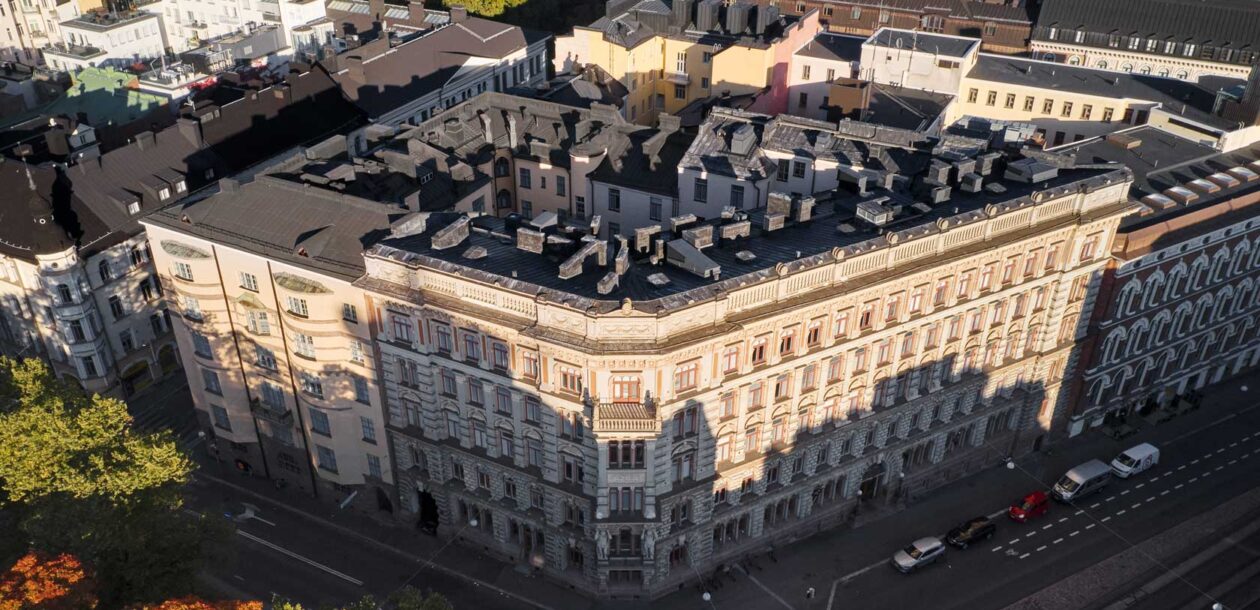 Historiallinen rakennus Helsingissä, jossa on yksityiskohtaisia arkkitehtonisia piirteitä. Rakennus sijaitsee kadun kulmassa, ja sen ympärillä on muita rakennuksia ja puita.