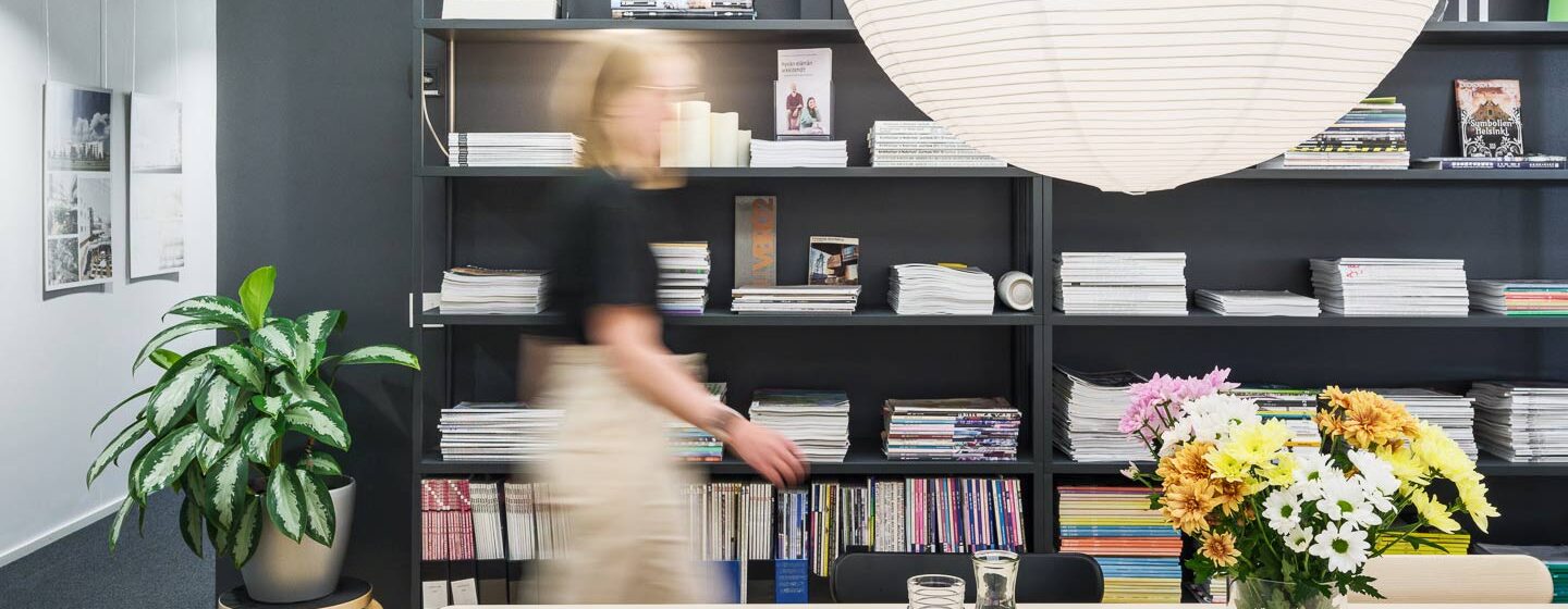 Moderni toimistotila, jossa katosta roikkuu suuri valkoinen paperilyhty. Etualalla on puinen pöytä, jolla on lasikannu, kaksi lasia, avoin kirja ja maljakko kukkia