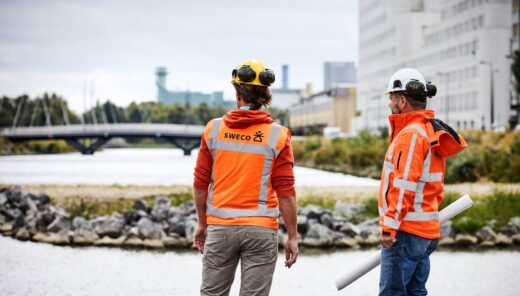 Kaksi rakennustyöntekijää, joilla on yllään oranssit huomiotakit, joissa on "SWECO"-logo ja teksti selässä. Toinen työntekijä pitelee kädessään rullattua piirustusta. He seisovat veden äärellä, taustalla näkyy silta ja rakennuksia.