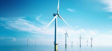 A group of offshore wind turbines standing in the ocean under a clear blue sky. The turbines are evenly spaced and extend into the distance