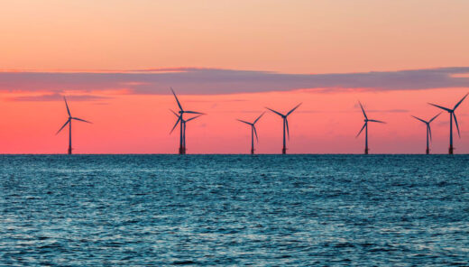 tuuliturbiineja merellä auringonlaskun aikaan. Taivas on oranssin ja vaaleanpunaisen sävyinen, ja tyyni vesi heijastaa näitä värejä.