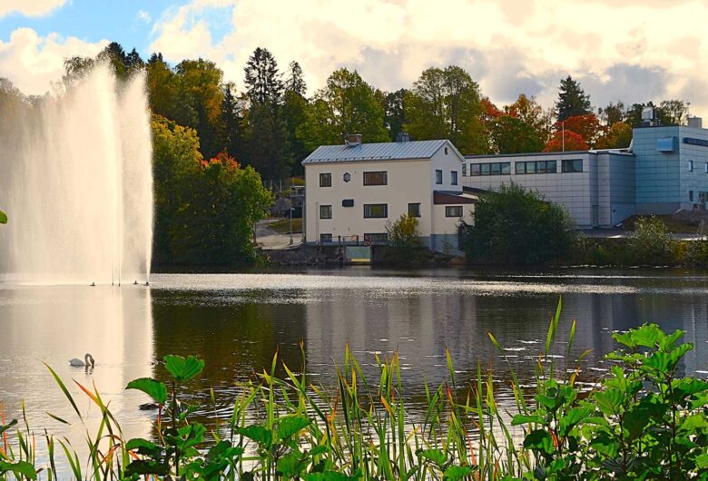 Järvimaisema, jossa näkyy suihkulähde, rakennuksia ja vehreää kasvillisuutta.