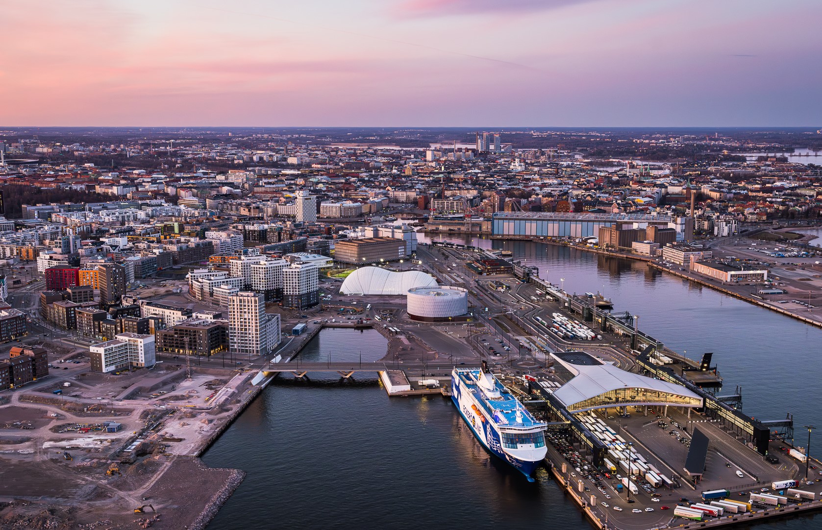 Ilmakuva Helsingin satama-alueesta, laiva, rakennuksia auringonlaskun aikaan.