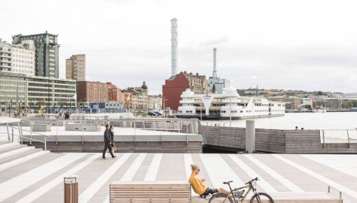 City waterfront with parked ferry, cyclist resting on bench, and pedestrians walking.