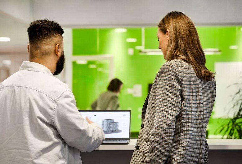 Two people discussing architectural plans on a laptop in a modern office with green walls.