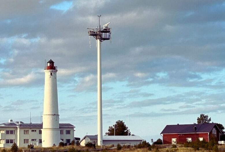 White lighthouse next to buildings under cloudy sky. Birds on tall pole. Scenery with trees and grass.