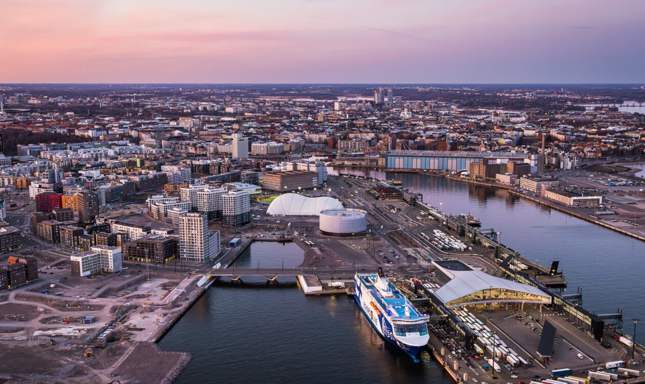 Ilmakuva Helsingin Jätkäsaaren alueesta auringonlaskun aikaan. Kuvassa näkyy satama-alue laivoineen, moderneja kerrostaloja ja rakennuksia, kaksi suurta valkoista rakennusta, useita pysäköityjä ajoneuvoja sekä kaupungin siluetti taustalla.