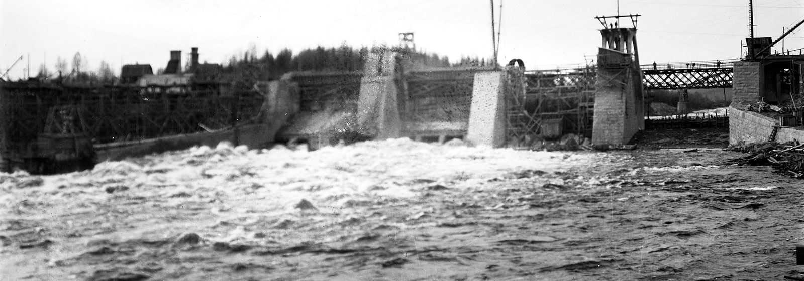 Mustavalkoinen kuva näyttää suuren padon, jonka läpi virtaa voimakas vesivirta. Taustalla näkyy rakennuksia ja metsää