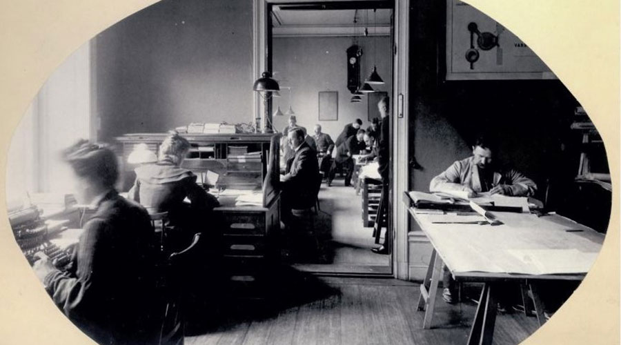 Mustavalkoinen valokuva Swecon toimistosta, jossa useita ihmisiä työskentelee vanhanaikaisten kirjoituskoneiden ja asiakirjojen parissa. Etualalla kolme henkilöä, taka-alalla useampia pöytien ääressä.