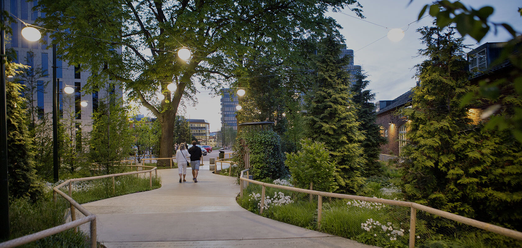 Kaupunkipuisto iltahämärässä, valaistu kävelytie, kaksi ihmistä kävelee polulla, ympärillä puita ja pensaita, taustalla korkeita rakennuksia