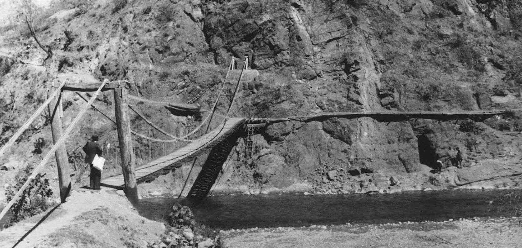 Vanha puu- ja köysirakenteinen riippusilta ylittää joen vuoristoisessa maisemassa. Henkilö seisoo sillan alkupäässä pitäen paperia kädessään.
