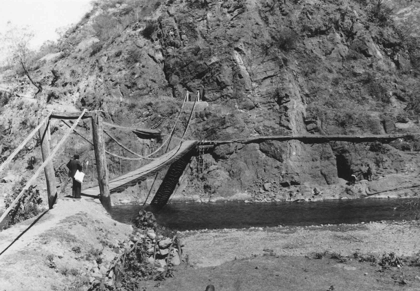 Vanha puu- ja köysirakenteinen riippusilta ylittää joen vuoristoisessa maisemassa. Henkilö seisoo sillan alkupäässä pitäen paperia kädessään.