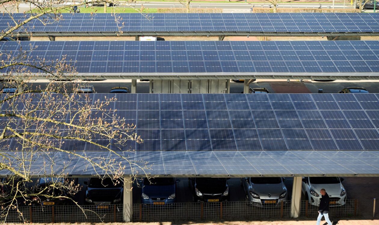 Pysäköintialue, jonka katoksina toimivat aurinkopaneelit. Useita autoja pysäköitynä katosten alla, etualalla kävelee henkilö. Taustalla on tie ja puita.