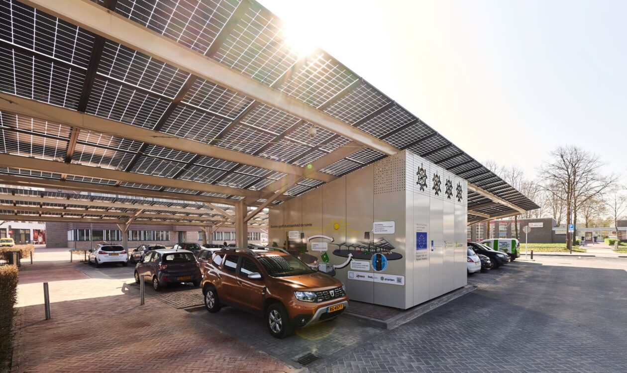 A parking area covered with solar panels, with several parked cars. In the foreground, a brown car; in the background, buildings and trees