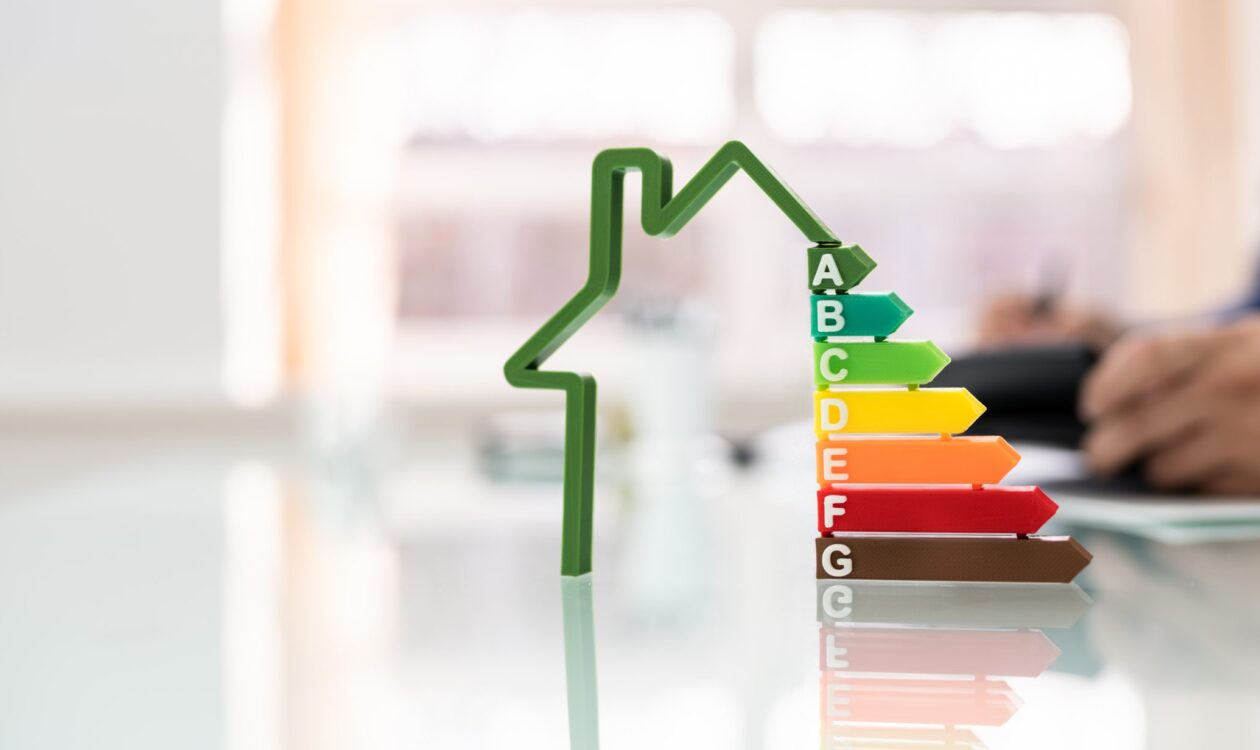 The image shows the energy efficiency rating, represented by a house-shaped symbol and colorful bars. The bars are labeled with letters from A to G, where A is green and G is red. In the background, a person is seen taking notes.