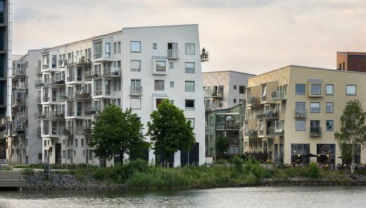 Valkoinen, vaalenakeltainen sekä punatiilinen kerrostalo veden äärellä, matalat, maksimissaan viisikerroksiset talot ja lasitetut, 2020-luvun tyyliset parvekkeet.