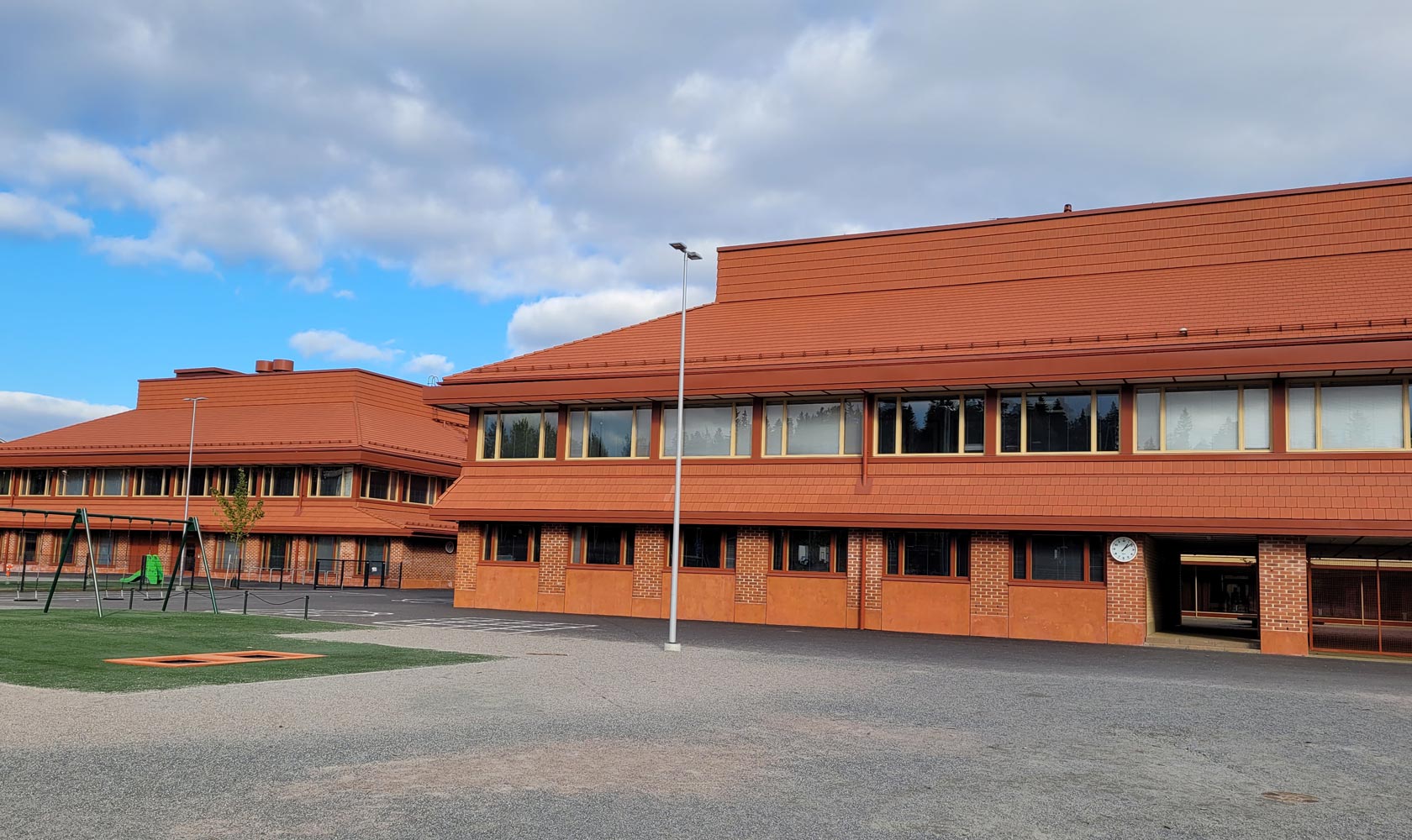 Modern school building with red brick facade, windows, playground, and cloudy sky.