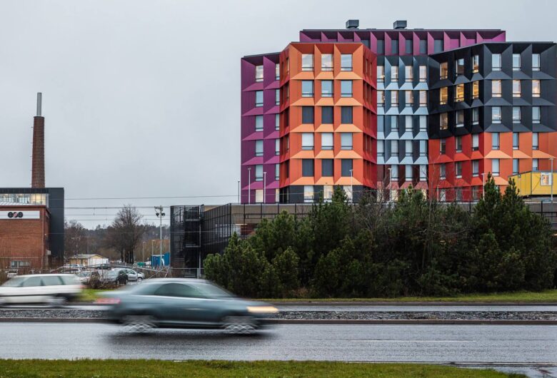 Värikäs rakennus Trivium ja liikkuvat autot sateisena päivänä, taustalla tiilikaupunki ja savupiippu.