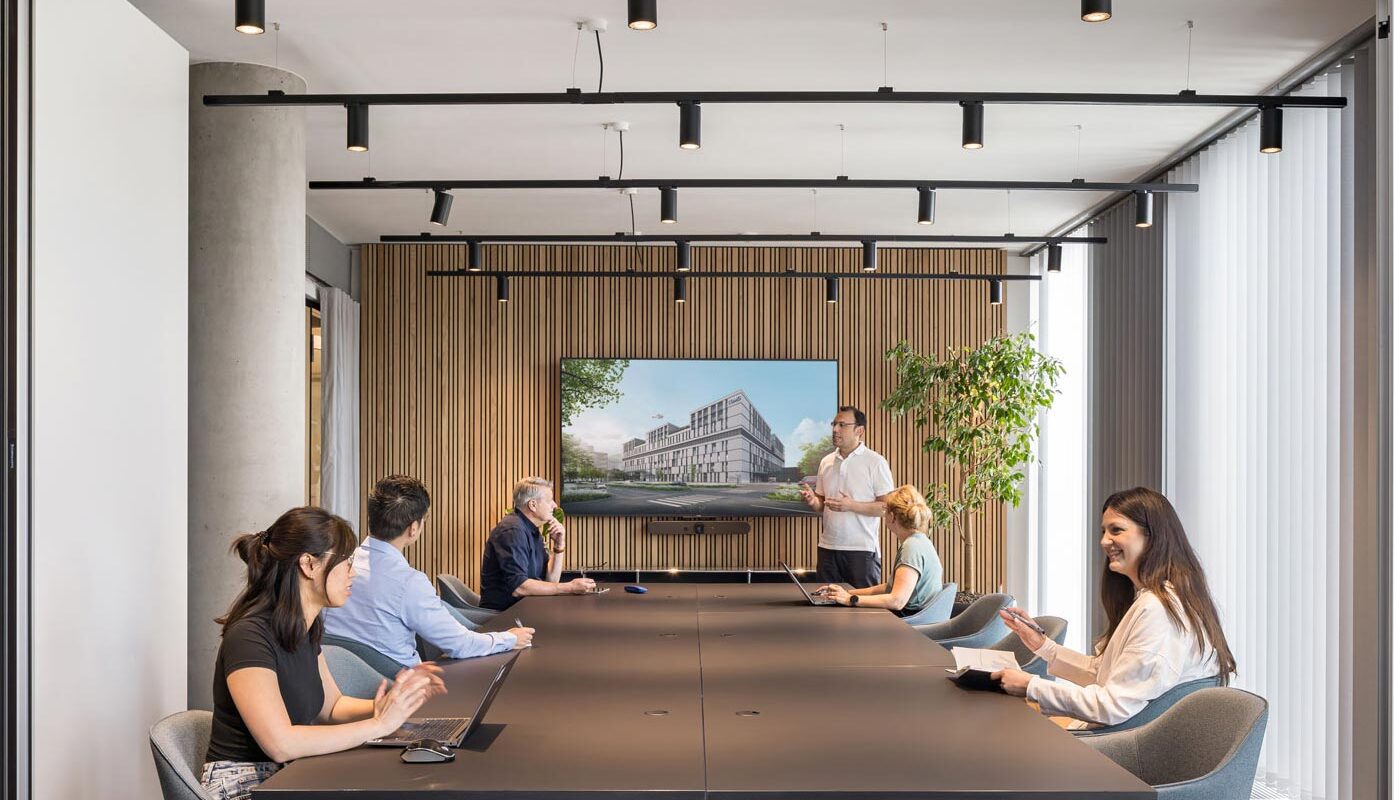 Viisi ihmistä kokoushuoneessa katsovat esitystä televisioruudulla.