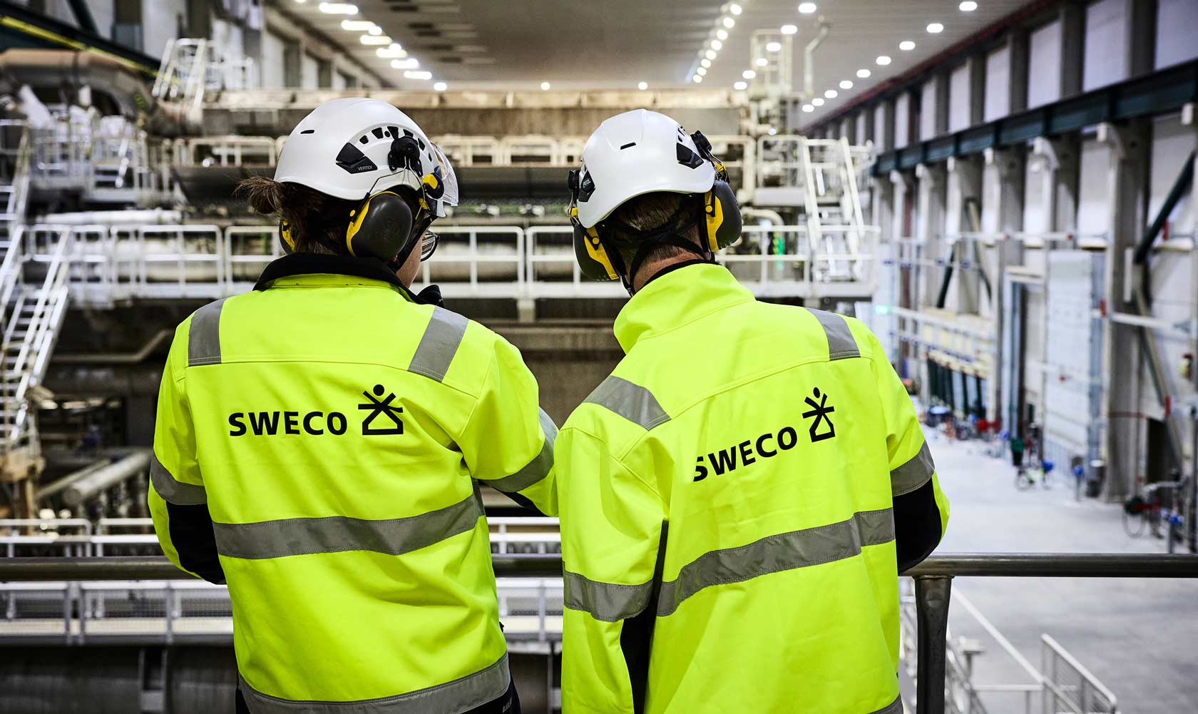 Kaksi Sweco-työntekijää keltaisissa vaatteissa ja kypärissä katsovat teollisuushallia.