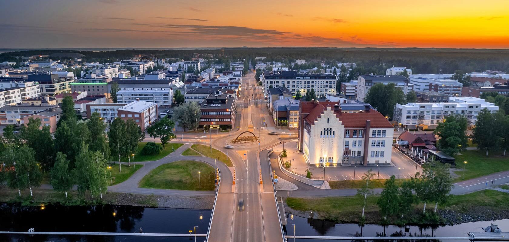 Ilmakuva kaupungista auringonlaskun aikaan, moderni kaupunkimaisema ja vesistö etualalla.