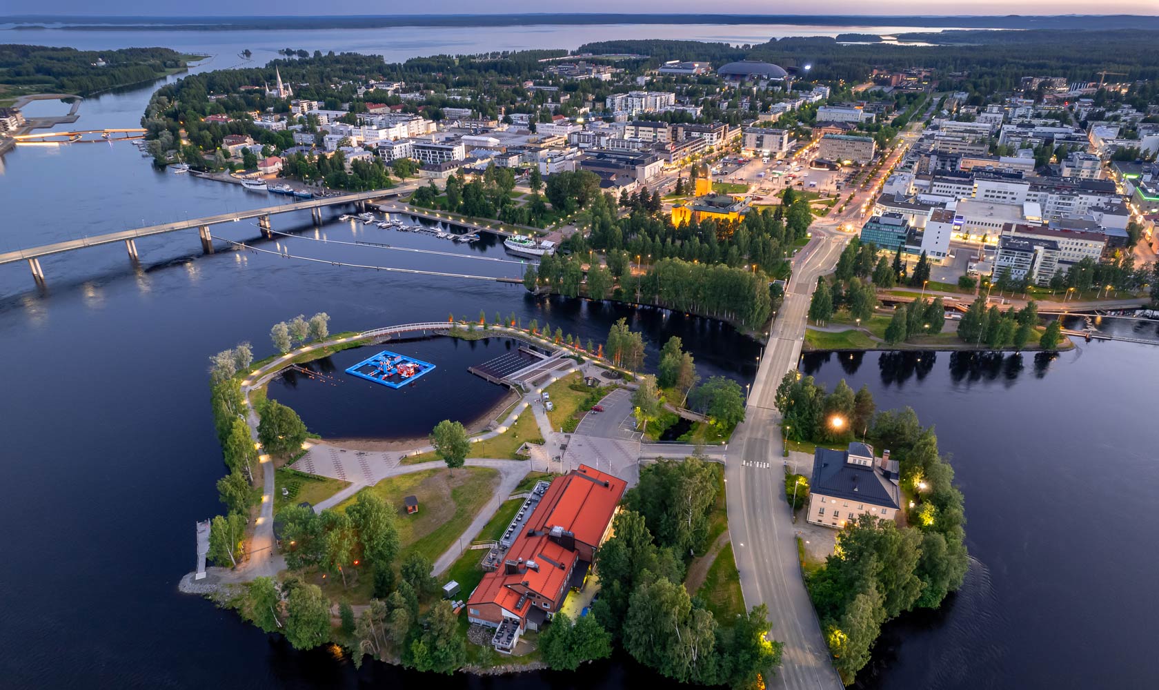 Ilmakuva Joensuusta, jossa näkyy kaupunki, siltoja ja vesistöjä iltahämärässä.